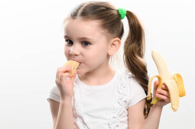 Patates cipsi yiyen ve elinde muz tutan küçük bir kız. Beyaz arka planda stüdyo fotoğrafı. Sağlıklı ve sağlıksız yiyecekler..