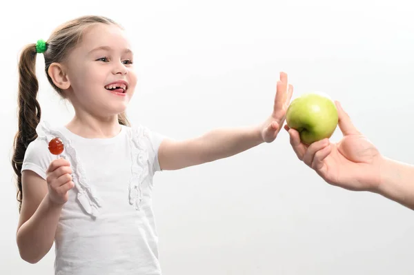 Little girl chooses a lollipop over an apple, sweets and fruits, choice between healthy and unhealthy food, white background and child.