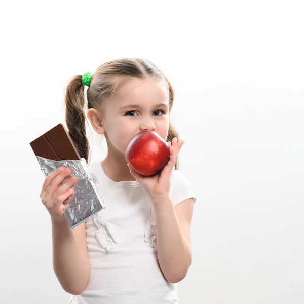 Little cute girl chooses apple over chocolate, preference of healthy food over unhealthy and unhealthy, child eats red apple on white background and holds chocolate in hand.