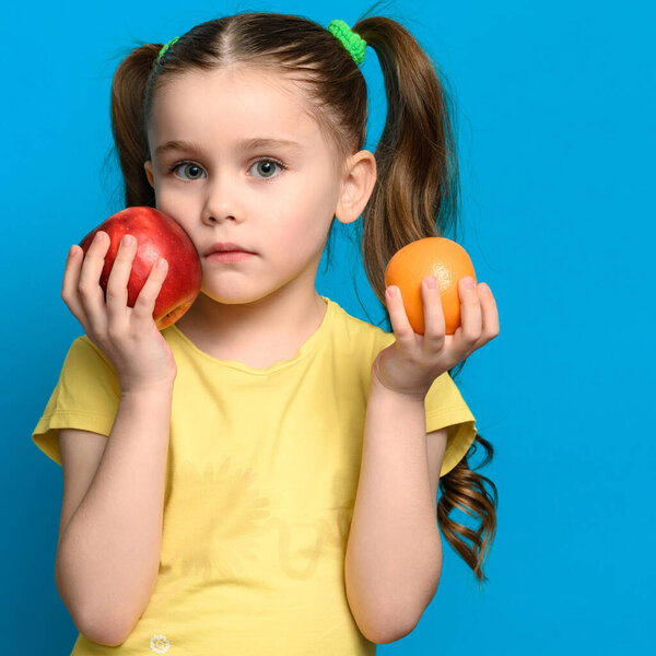 On a blue background, the girl holds an apple and a tangerine near her cheek, fruits for baby food.