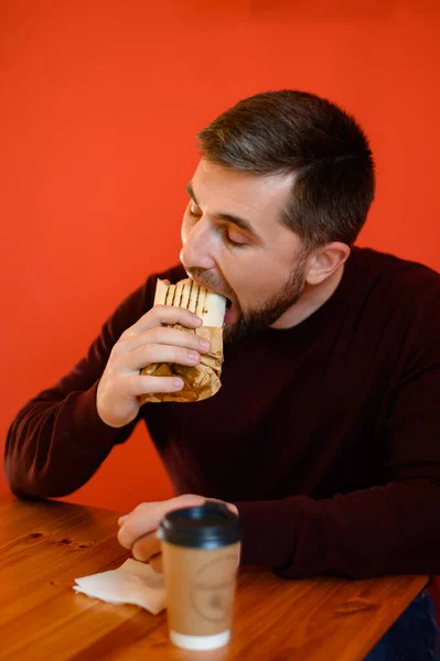 男はファーストフードのレストランに座って 赤い壁の背景に対してショールマルタをかむ 迅速な軽食のためのショールマルタ — ストック写真