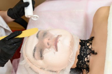 Hastanın yüzü gazlı bezle kaplıdır, güzellik uzmanı kızın yüzüne spatulayla krem sürer, kuru ciltle ilgilenir..