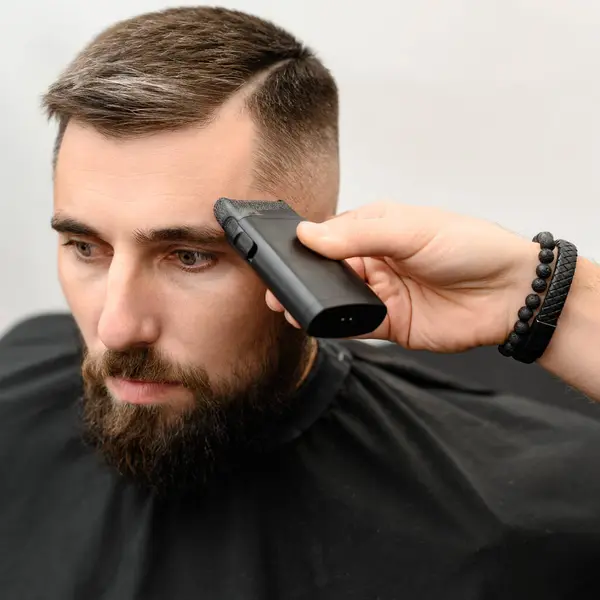 Barber shaves the temple with a cordless trimmer during a short haircut on the sides of the head.
