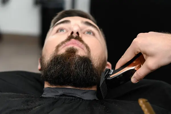 Cutting a gentleman beard in a barbershop with a clipper. Shortening the length of the beard from the sides by the master for the client.