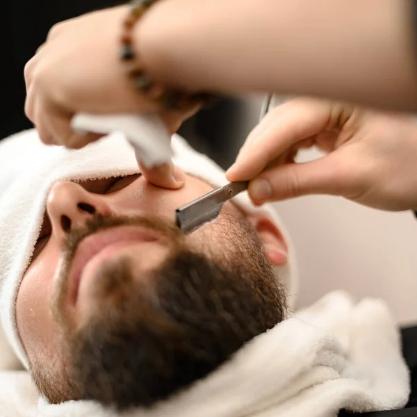 A barber shaves the cheek of a bearded customer with a dangerous razor. Shaving the contour of the beard for the correct shape.