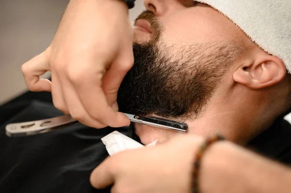 A barber shaves the neck of a bearded customer with a dangerous razor. Shaving the contour of the beard for the correct shape.
