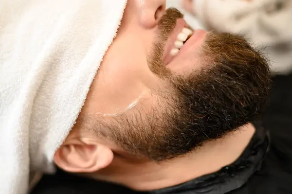 Shaving gel on the face of a smiling bearded man in a barbershop with a towel over his eyes