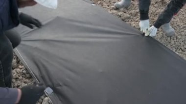 Bir adam plastik siyah kazığı yerde tutmak için tarım ipliğine saplıyor. Bahçıvanlar tarlada ve bahçede çalışıyor..