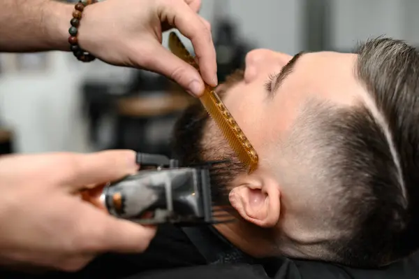 Barber combs beard with comb while cutting and shaving Caucasian man