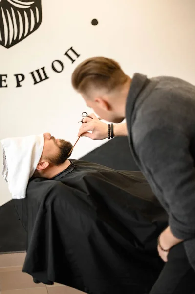 Barber combs beard with comb while cutting and shaving Caucasian man