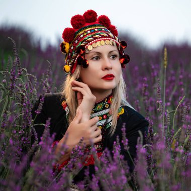 A girl in a chelsea headdress and a black dress decorated with red embroidery sits between lavender bushes, Ukrainian traditional and national clothing.