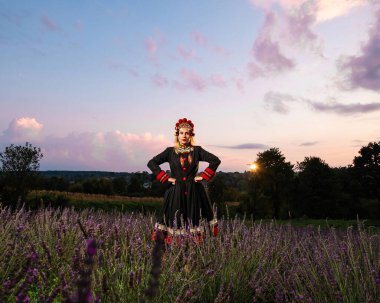 A girl in a chelsea headdress and a black dress decorated with red embroidery stands between lavender bushes.