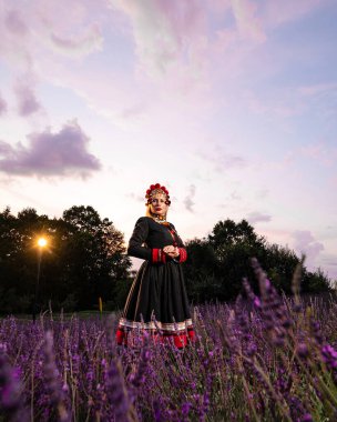 A girl in a chelsea headdress and a black dress decorated with red embroidery stands between lavender bushes.