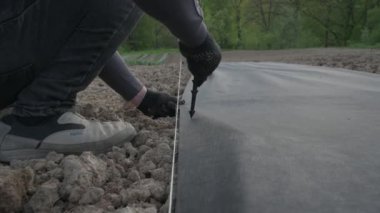 Bir adam plastik siyah kazığı yerde tutmak için tarım ipliğine saplıyor. Bahçıvanlar tarlada ve bahçede çalışıyor..