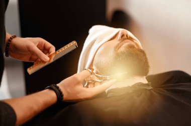 Bir berber stilisti, yüzü havluyla, makasla kaplı beyaz bir adamın sakalını keser.