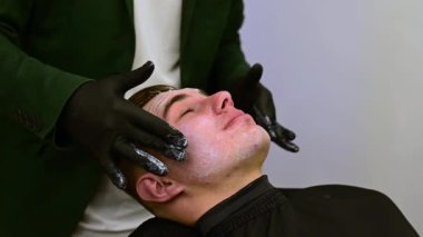 Siyah eldivenli bir berber müşterinin yüzüne nemlendirici beyaz krem sürer. Bir güzellik uzmanı beyaz bir maske takar ve bir erkeğin tenine vitamin kompleksi uygular..