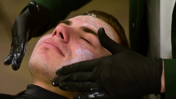 一个戴着黑色手套的理发师在客户的脸上涂上保湿的白霜 一位美容师在一个面临慢动作视频的男人的皮肤上涂上带有维生素复合体的白色面膜 — 图库视频影像