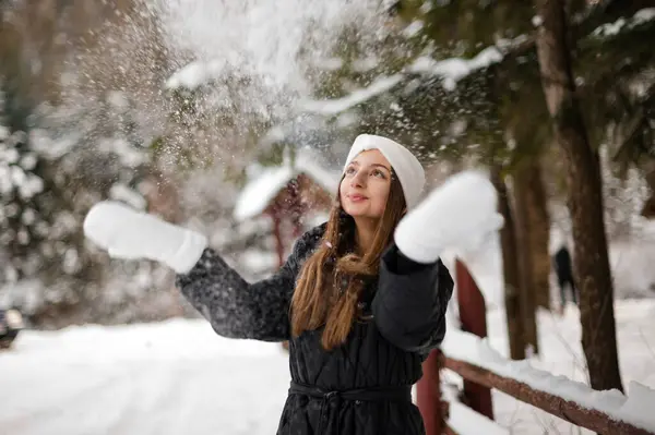 Portrait of a girl in winter clothes with white gloves and a white headband near a wooden fence and forest. The girl scatters snow from her hands, white gloves and a black jacket.