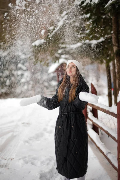 Portrait of a girl in winter clothes with white gloves and a white headband near a wooden fence and forest. The girl scatters snow from her hands, white gloves and a black coat.