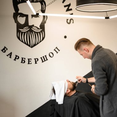 Bir berber stilisti, yüzü havluyla, makasla kaplı beyaz bir adamın sakalını keser.