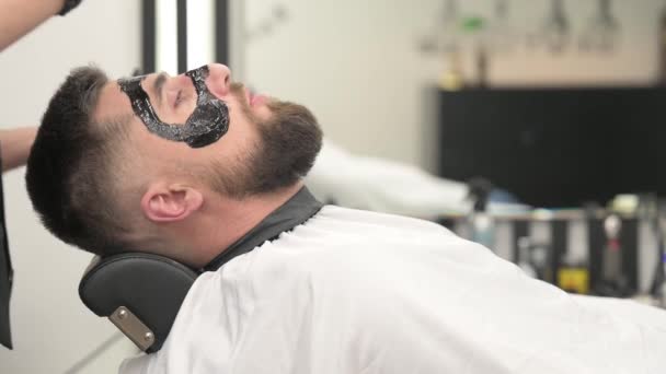 バーバーは シェービングしている間 マンションの顔に黒い保湿マスクを塗ります 皮膚科医は ひげを被った患者の顔に黒いマスクを塗り 毛穴をクリアし 4Kビデオをしわにします — ストック動画