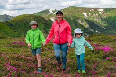 A mother with her son and daughter are walking among rhododendron bushes against the background of the majesty of the Carpathian Mountains, active summer vacation with children. clipart