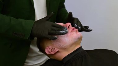 Siyah eldivenli bir berber müşterinin yüzüne nemlendirici beyaz krem sürer. Bir güzellik uzmanı beyaz bir maske takar ve bir erkeğin tenine vitamin kompleksi uygular..