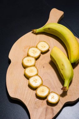Taze muzlar, bir bütün ve bir dilimlenmiş. Koyu arkaplana karşı ahşap bir kesme tahtasına dizilmiş. Dilimler düzgünce yerleştirilmiş, meyvenin dokusunu ve rengini gösteriyor..