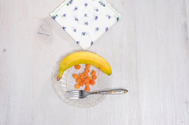 Tek bir muz ve bir avuç sarı ahududu, bir çatalın yanındaki cam bir tabağa yerleştirilir. Hafif ahşap masanın üzerinde desenli bir peçete duruyor..