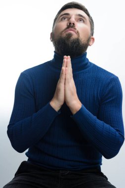 Mavi boğazlı kazaklı sakallı bir adam elleri dua ederken sıkıca oturup beyaz bir arka plana karşı umut dolu bir ifadeyle yukarıya bakar..