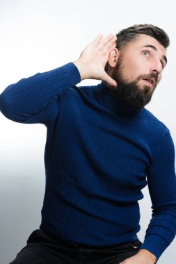 Mavi balıkçı yaka kazak ve koyu pantolon giyen sakallı bir adam elini kulağına dayayıp arka planda hafif bir ses duymaya çalışıyor..