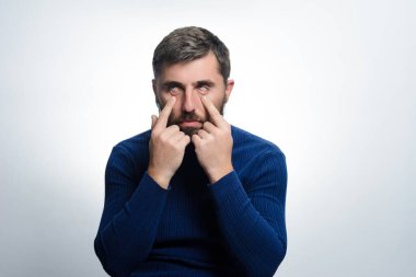 Mavi kazak giyen sakallı bir adam parmaklarını gözlerinin etrafındaki deriye dokunmak için kullanır. Açık gri bir arkaplanda kameraya bakıyor..