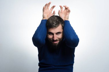 Mavi kazaklı sakallı bir adam açık gri bir arka plana karşı elleri havada şakacı hareketler yapar. Yaramaz ve tasasız bir ifade yaratır..