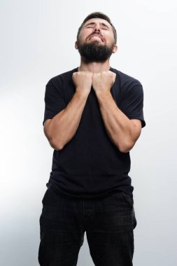 Siyah gömlekli ve siyah pantolonlu sakallı bir adam, açık arkaplan karşısında, ellerini göğsüne tutarak, elleri kapalı ve yüzü bükülmüş bir şekilde, saf bir duygu sergiliyor..