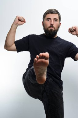 Koyu renk gömlekli ve kot pantolonlu sakallı bir adam tekme atıyor. Yumruklarını güçlü ve dinamik bir pozda hafif bir arkaplana doğru kaldırırken ayağını kameraya doğru kaldırdı..