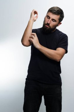 Siyah gömlek ve siyah pantolon giyen sakallı bir adam kolunu düz beyaz bir arka plana sürterek kaşır. Rahatsızlık ya da rahatsızlık hissediyor gibi görünüyor..