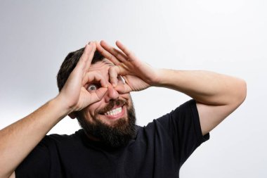 Şakacı bir ifadeye sahip sakallı bir adam ellerini kullanarak gözlerinin etrafında dürbün oluşturur, hafif bir arka planda geniş bir gülümsemeyle. Onun şakacı jesti bir neşe ve rahatlık hissi veriyor..