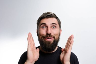 Sakallı bir adam şaşkınlığını geniş gözlerle gösterir ve hafif bir arka plana karşı ellerini kaldırır. Neşeli ifadesi şaşkınlık ve heyecan veriyor..