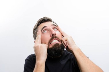 Beyaz bir geçmişi olan sakallı bir adam endişeli ve stresli bir ifadeyle yukarı bakar. Parmaklarıyla şakaklarına dokunuyor. Gerginlik ya da baş ağrısı olduğunu gösteriyor..
