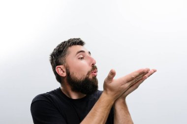 Siyah gömlekli sakallı bir adam beyaz bir arka plana öpücük yolluyor. Ellerini sanki havaya narin bir şey yolluyormuş gibi tutuyor..