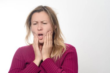 Bir kadın çenesini iki eliyle tutar, yüzü rahatsızlık ve endişe gösterir. Dişlerinde ya da çene bölgesinde sade bir arka planda ağrı ya da rahatsızlık hissediyor gibi görünüyor..