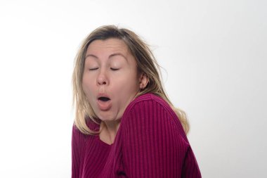 Omuz hizasında sarı saçlı, kestane rengi kazak giyen bir kadın, ağzı açık ve gözleri kapalı beyaz bir arka plana karşı gösteriliyor..