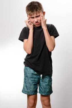 Siyah tişörtlü ve desenli şortlu bir çocuk, elleri şakaklarına değmiş bir şekilde baş ağrısı çekiyor. Rahatsız ve stresli görünüyor..