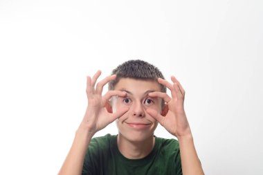 Genç bir çocuk komik bir yüz ifadesi takınarak elleriyle gözlerini kapatıyor. Yeşil bir gömlek giyiyor ve neşeli bir ifadesi var. Arkaplan düz beyaz.