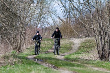 Bir çift birey, yapraksız ağaçlarla çevrili toprak bir patika boyunca bisiklet sürüyor. Sahne, ilkbaharın başlarında bir dinginlik ve açık hava aktivitesi hissi uyandırıyor..