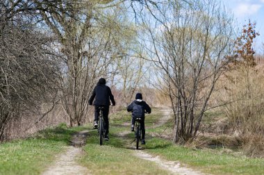 Ağaçlar ve yeşilliklerle çevrili iki kişinin toprak bir patika boyunca bisiklet sürdüğü huzurlu bir manzara. Barışçıl ortam, rahatlatıcı bir açık hava aktivitesi olduğunu gösteriyor..