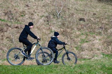 Dağ bisikleti süren iki genç insan çimenli bir yolda bisiklet sürüyor. Arka planda hafif eğimli bir yamaç var. Sahne güneşli bir gün ve açık hava eğlencesi olduğunu gösteriyor..