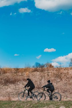 İki siyah giysili birey, açık mavi gökyüzü ve kuru, kahverengi çimenlerin arka planında yan yana dönerler. Sakin bir açık hava sahnesi.