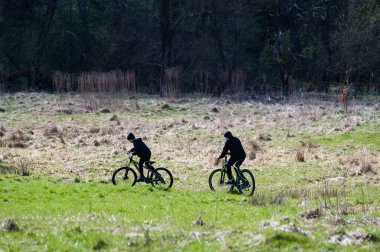 Çimenli bir alanda bisiklet süren iki kişinin sakin bir sahnesi. Güneşli çayır, arka plandaki karanlık ağaç siluetleriyle çelişiyor ve sakin bir atmosfer yaratıyor..