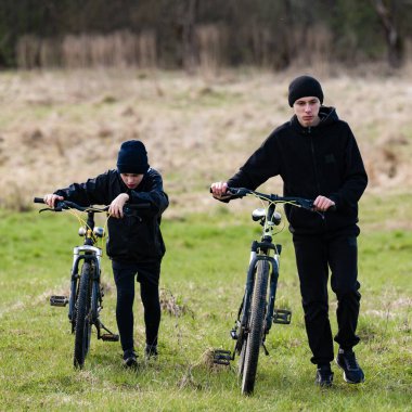 Siyahlar giymiş iki genç, gelişigüzel bisikletleriyle canlı yeşil bir alanda yürüyorlar. Ortam huzurlu ve güneşli..